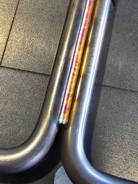 Lasergeschweißtes Rundmaterial Verbindung zweier metallischer Rohre auf einem schwarzen Bodenbelag.